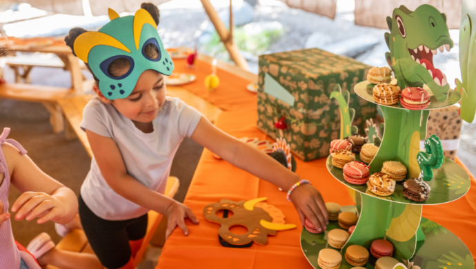 Niño con máscara de dinosaurio tomando galletas de la mesa de fiesta