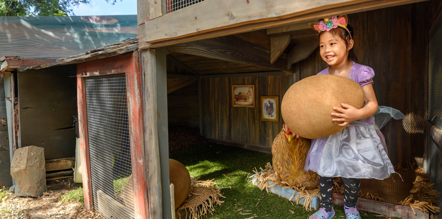 girl with bow in hair holds a giant egg in a chicken coop